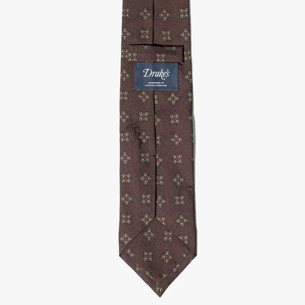 A brown silk necktie with a repeating geometric cross pattern in beige and gold, featuring a blue brand label that reads 'Drake's'.