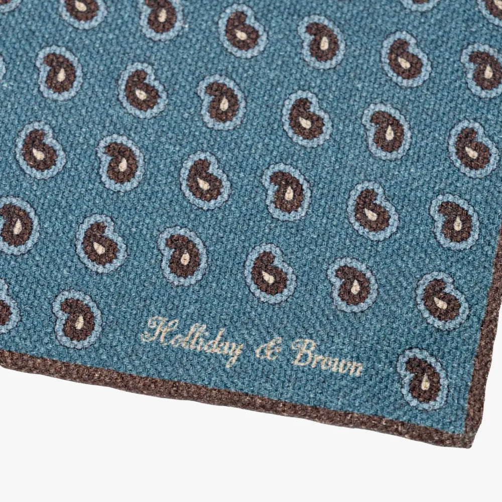 Close-up of the Holliday & Brown blue paisley pocket square corner highlighting the border, paisley print details, and brand logo.