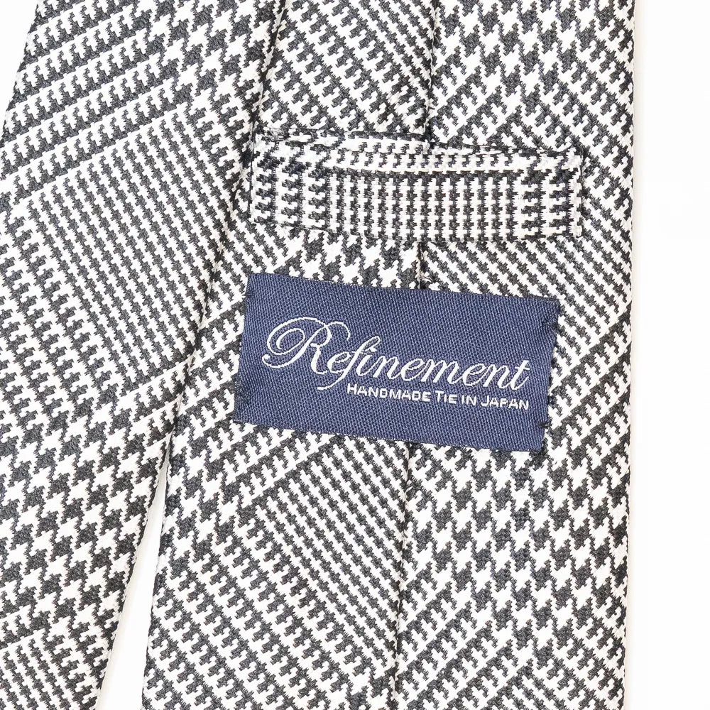 Close-up of the Refinement Prince of Wales neck tie showing the textured silk weave and brand label detail.