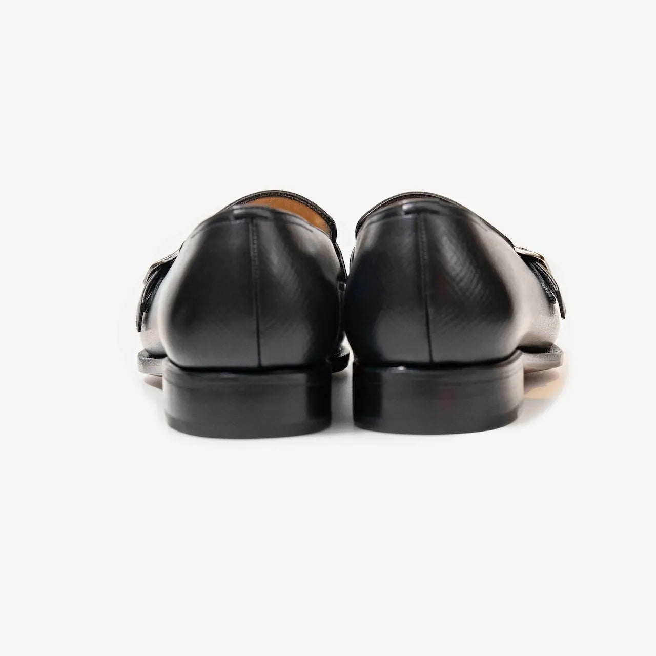 Rear view highlighting the heel shape and fine stitching of the black hatch grain buckled loafers.